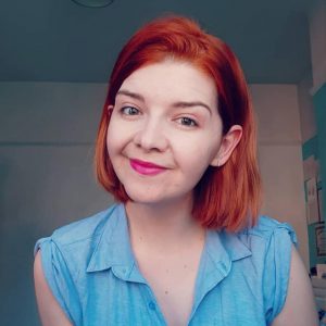 Gemma Louise Walsh has short red hair and is wearing a blue shirt and pink lipstick.