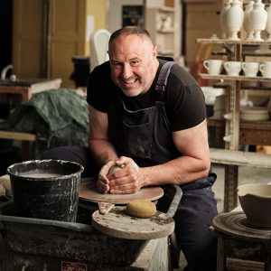 Keith Brymer-Jones sits at a potters wheel sculpting clay. He is wearing a black t-shirt and navy blue overalls.