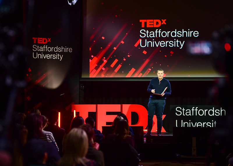 Event presenter Martin Tideswell is standing on the stage, with ted x Staffordshire University branding behind him. He is holding a red book, and wearing a navy blue jumper and trousers.