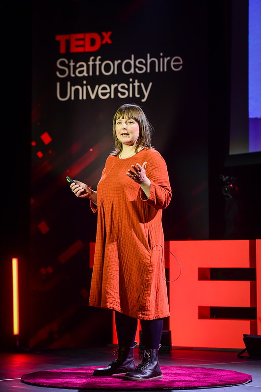 Professor Anna Francis delivers her talk from the Ted x Staffordshire University stage. She is wearing an orange dress and brown boots.