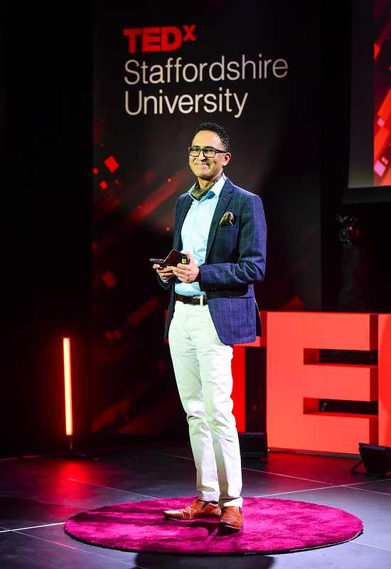 Professor Raheel Nawaz smiles out to the audience from the ted x Staffordshire University stage. He is wearing a Blue suit jacket and shirt with cream trousers. He has dark green ascot and matching pocket square.