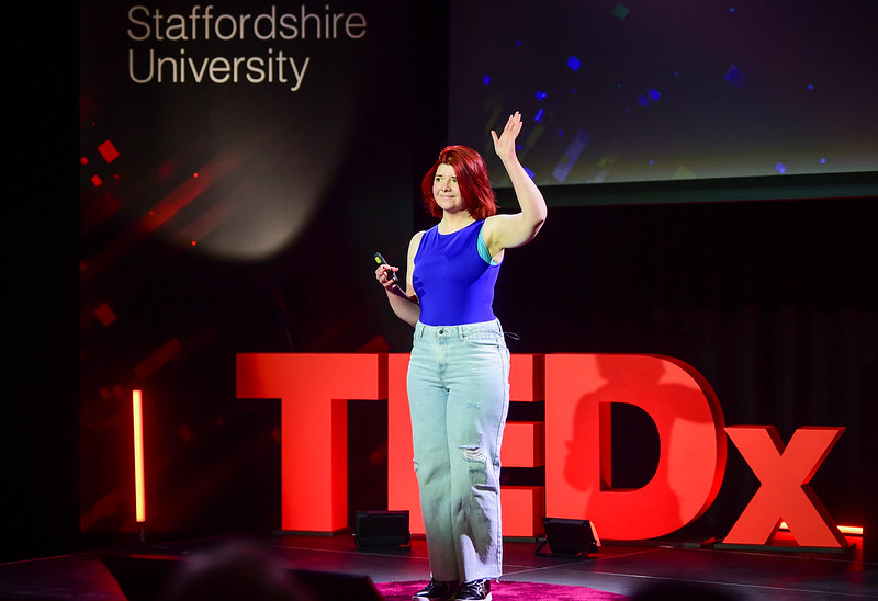 Gemma Louise Walsh is waving to the audience from the ted x Staffordshire University stage. She has short red hair and is wearing a blue vest top and jeans.