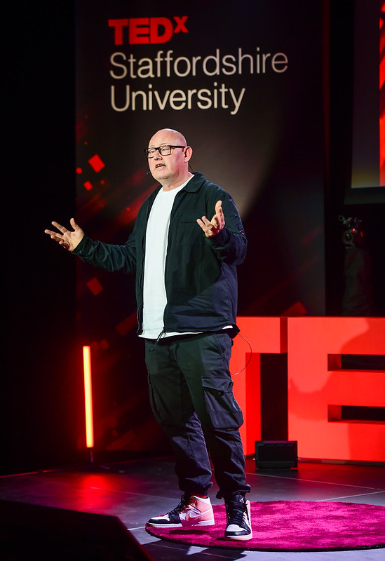 Darren Murinas is standing on a red rug on the ted x Staffordshire University stage, arms and hands outstretched. He is wearing Nike Dunk High top trainers, black glasses, a white t-shirt, a black jacket and trousers.