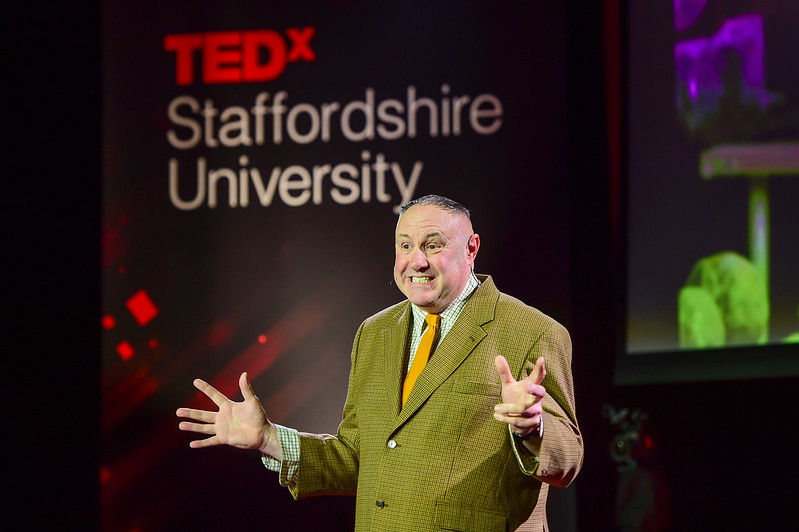 Speaker Keith Bryner Jones standing on stage, with his arms and hands out stretched. He is wearing a tattersall checked shirt, mustard yellow tie and green tweed blazer. He is smiling.